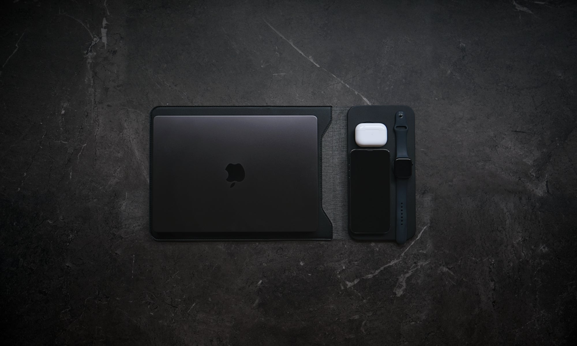 Black laptop and phone on a dark textured surface.NODE. Minimalist Black Laptop Sleeve with Apple Magsafe compatible charging docks for iPhone, Airpods and Apple Watch.