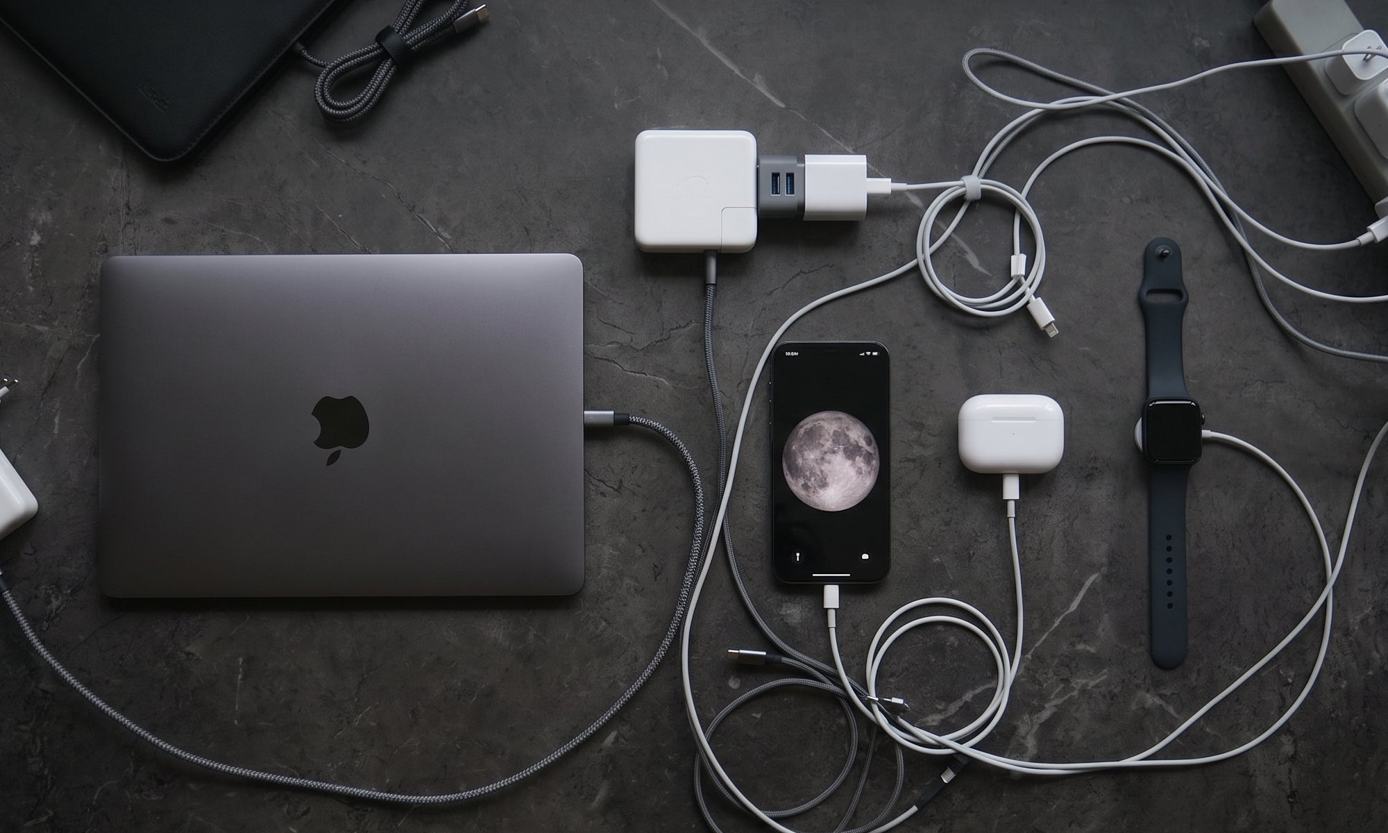A mess of Laptop, smartphone, smartwatch, and charging cables on a dark surface.