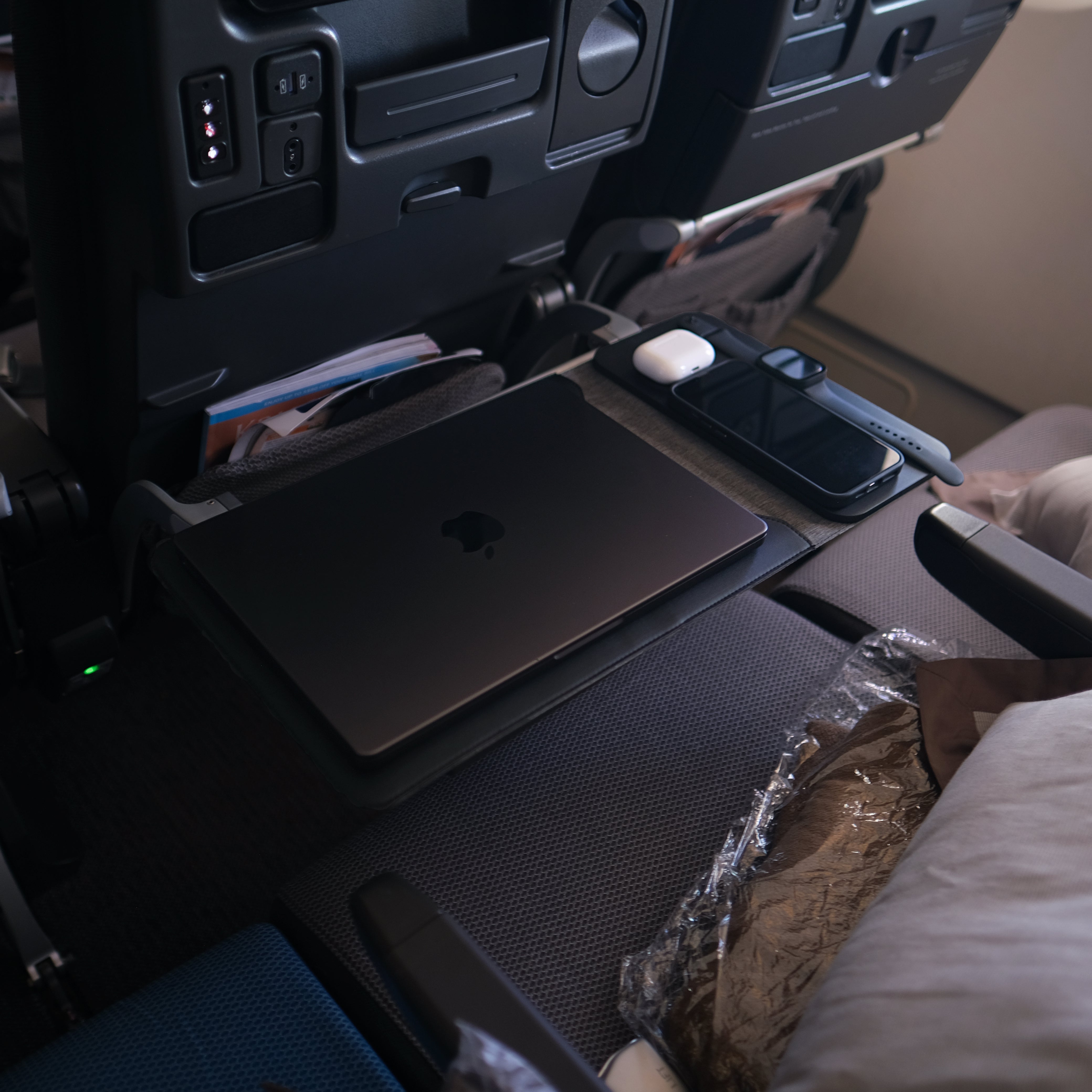 NODE. Minimalist Black Laptop Sleeve with Apple Magsafe compatible charging docks for iPhone, Airpods and Apple Watch. on a tray table in an Economy Class Singapore Airlines airplane.