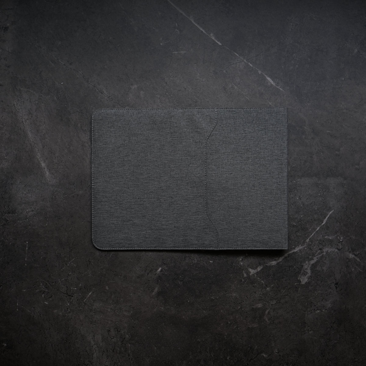 Dark gray textured card on a dark stone background.NODE. Minimalist Black Laptop Sleeve with Apple Magsafe compatible charging docks for iPhone, Airpods and Apple Watch.
